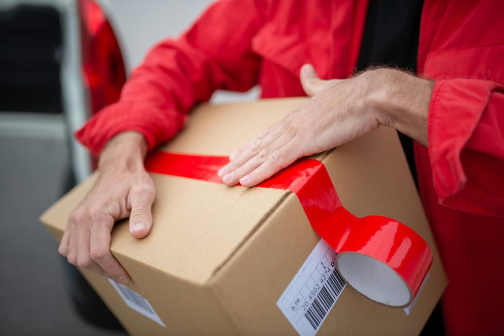 A courier in red uniform sealing a cardboard package with red tape for delivery.